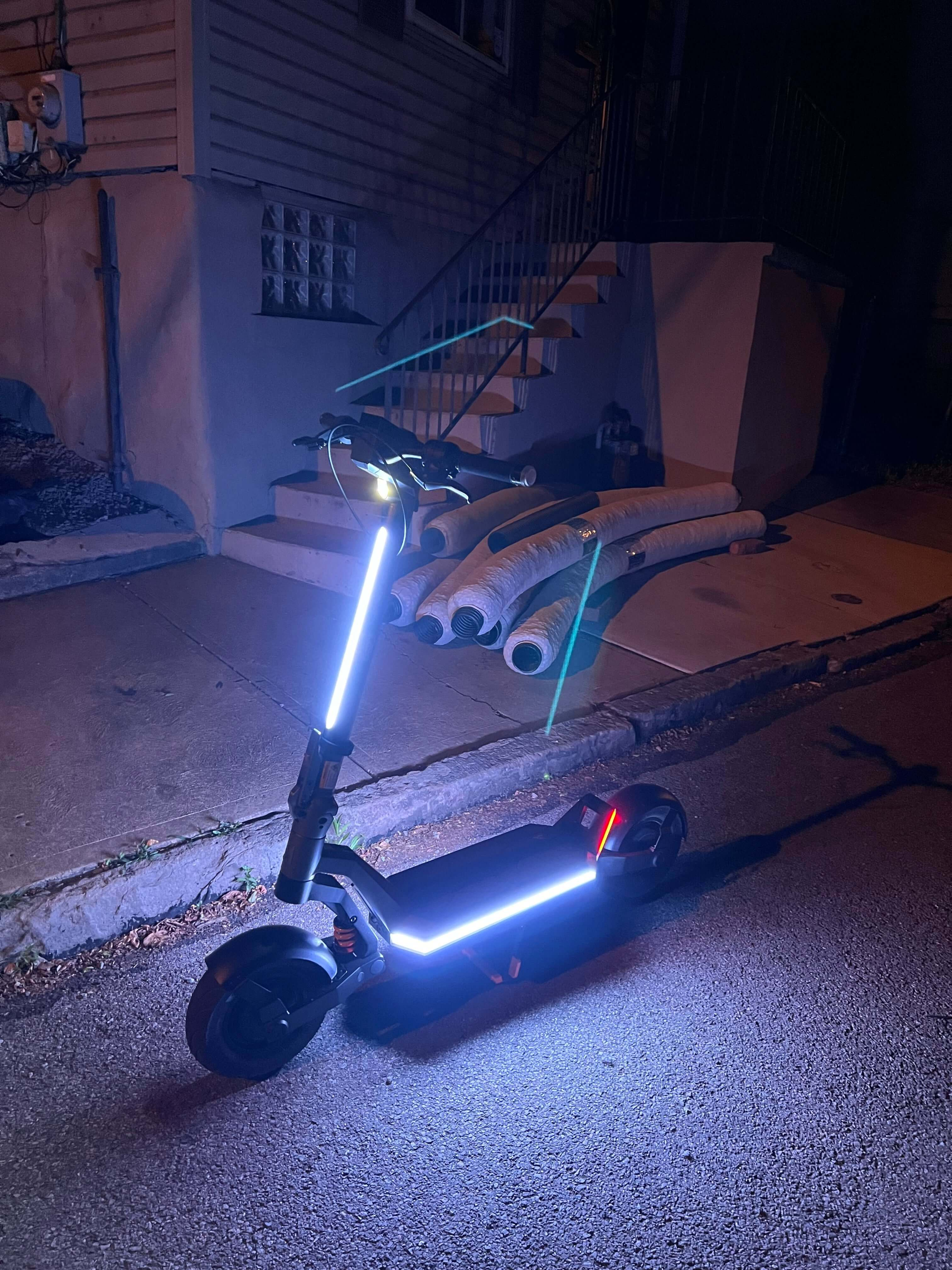Electric scooter with illuminated stem and deck lights parked on a street at night.