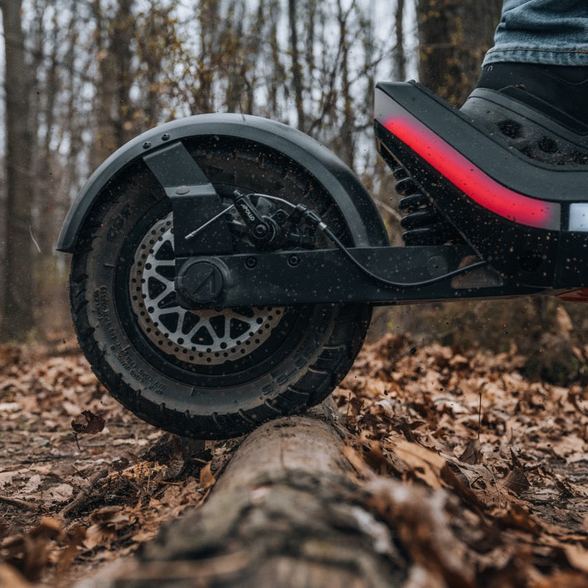 Close-up of a black electric scooter wheel and fender on a leaf-covered log in a wooded outdoor setting, with part of a person's leg and shoe visible on the deck.