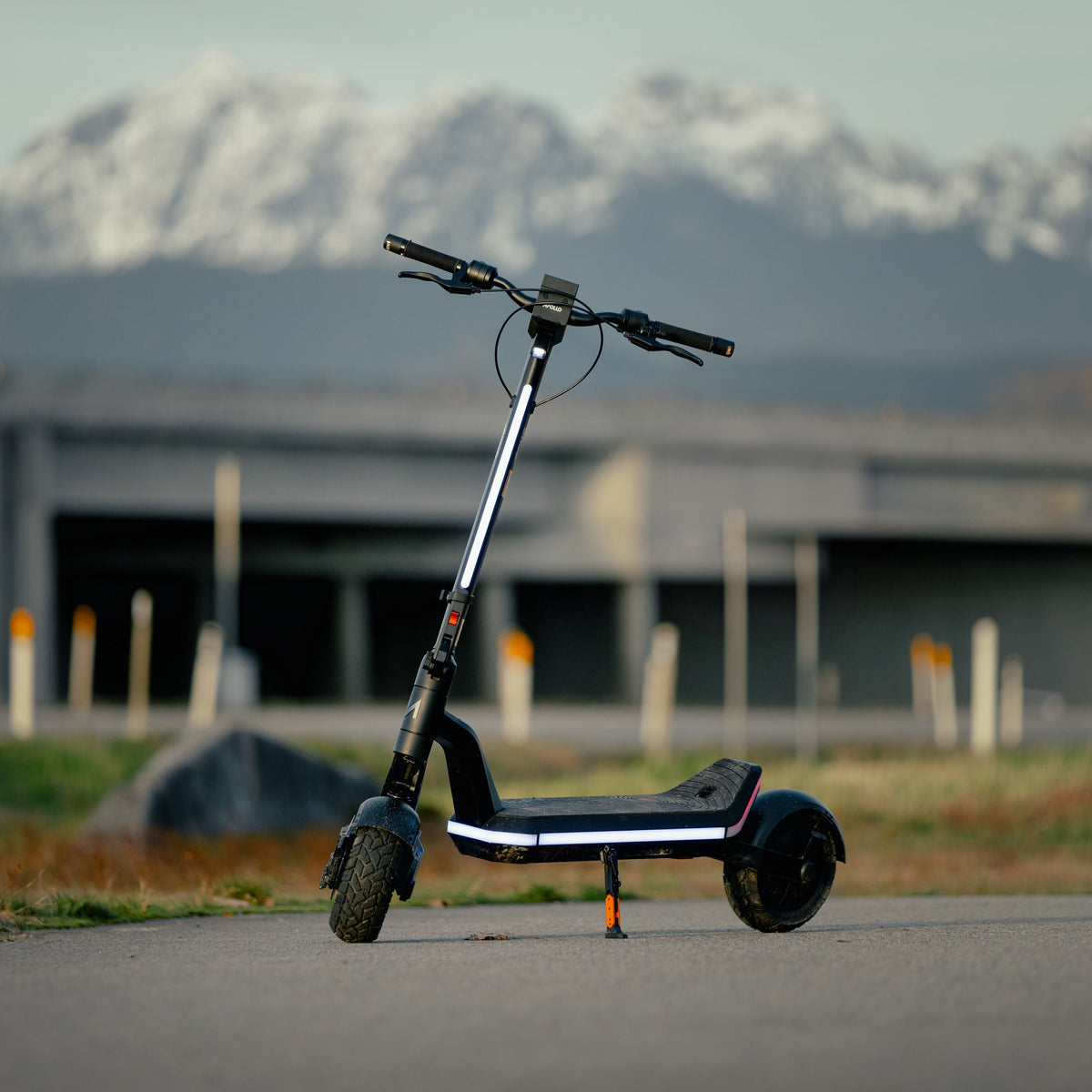 A black and silver electric scooter is parked on an asphalt road with a blurred background of a concrete structure and snow-capped mountains under a pale sky.