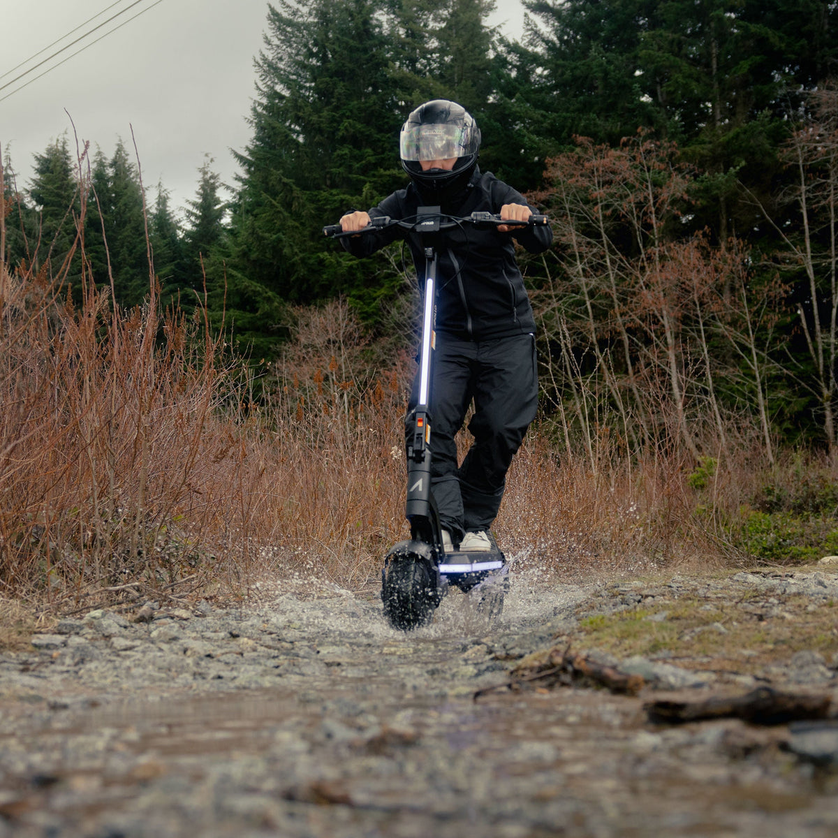 A person riding an electric scooter on a rocky dirt path outdoors, with dry brush and evergreen trees in the background.
