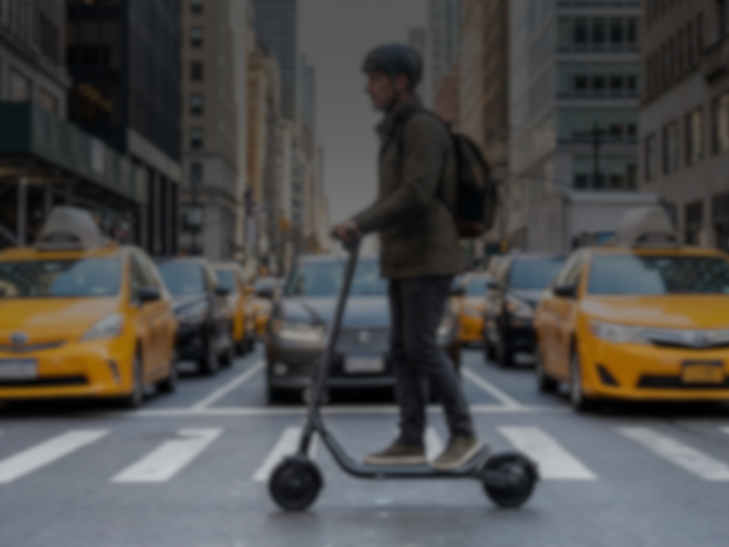 Person riding an electric scooter on a city street with yellow taxis in the background